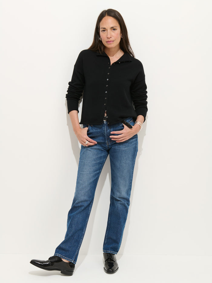 A woman with long dark hair stands against a white background, hands in her pockets, wearing the Taylor Cardigan in Cotton Cashmere, blue jeans, and black shoes. She appears relaxed.