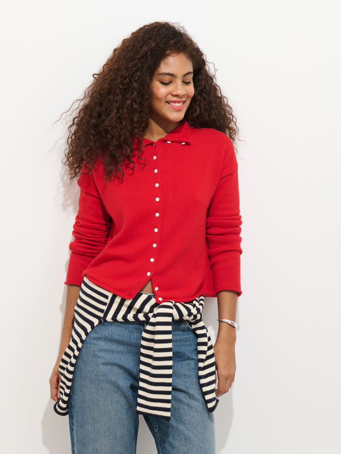 A woman with curly hair smiles in the Taylor Cardigan In Cotton Cashmere—red in color—worn over blue jeans, with a black-and-white striped sweater tied at her waist, standing against a plain white background.