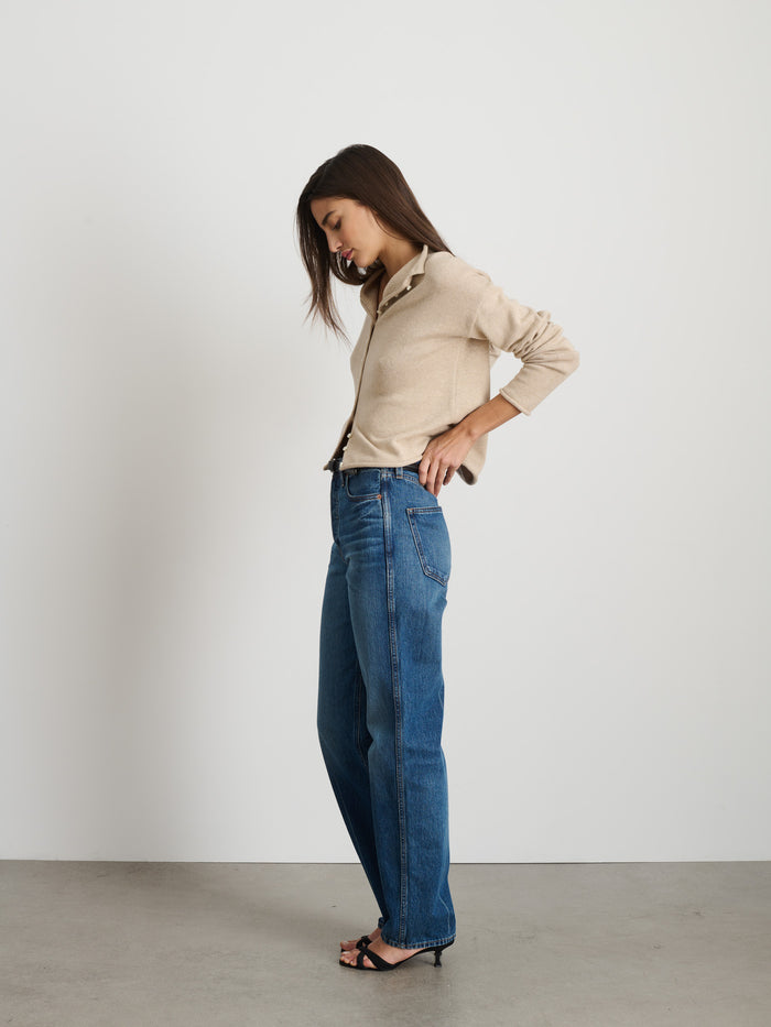 A woman with long brown hair stands in profile against a white wall, wearing the Taylor Cardigan In Cotton Cashmere, blue high-waisted jeans, and black heeled sandals. She looks down slightly with one hand on her hip.