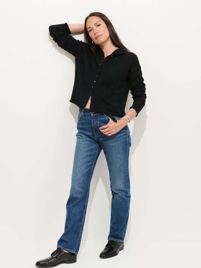 A woman stands against a white background wearing the Taylor Cardigan in Cotton Cashmere, blue jeans, and black shoes. With one hand on her head and the other in her pocket, she looks confidently at the camera.