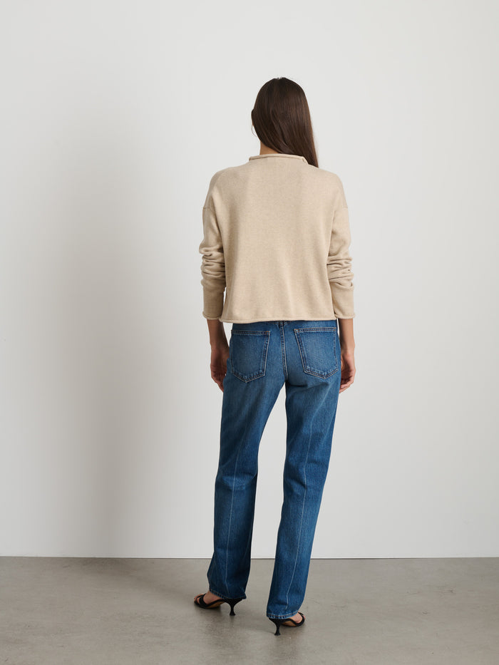A woman, seen from behind against a plain wall and concrete floor, wears the Taylor Cardigan in Cotton Cashmere, paired with blue jeans and black heeled sandals.