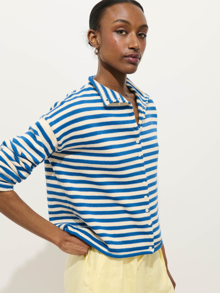 A woman wearing the Taylor Striped Cardigan in cotton-cashmere, featuring blue and white stripes and an oversized boxy fit, pairs it with light yellow pants. She poses confidently against a plain light background, hand on her hip.