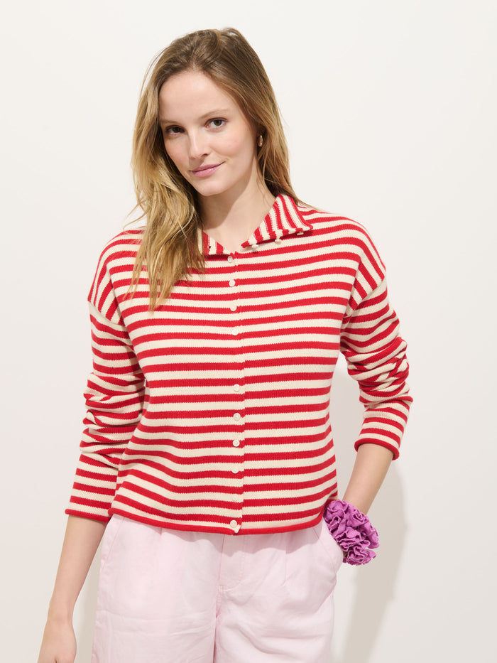 A woman with long blonde hair stands against a white background, wearing the Taylor Striped Cardigan in Cotton Cashmere with red and white stripes, light pink pants, and a purple scrunchie on her wrist.