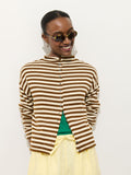 A smiling person wears tortoiseshell sunglasses, light yellow pants, and the Taylor Striped Cardigan in Cotton Cashmere with Troca shell buttons over a green top, standing in an oversized boxy fit against a plain white background.