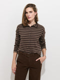 A person stands against a white wall, wearing the Taylor Striped Cardigan in Cotton Cashmere—an oversized brown and beige striped long-sleeve sweater with Troca shell buttons—tucked into high-waisted brown pants, with one hand in a pocket.