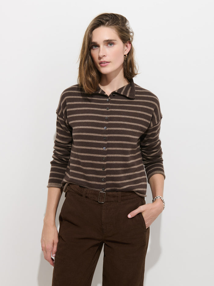A person stands against a white wall, wearing the Taylor Striped Cardigan in Cotton Cashmere—an oversized brown and beige striped long-sleeve sweater with Troca shell buttons—tucked into high-waisted brown pants, with one hand in a pocket.