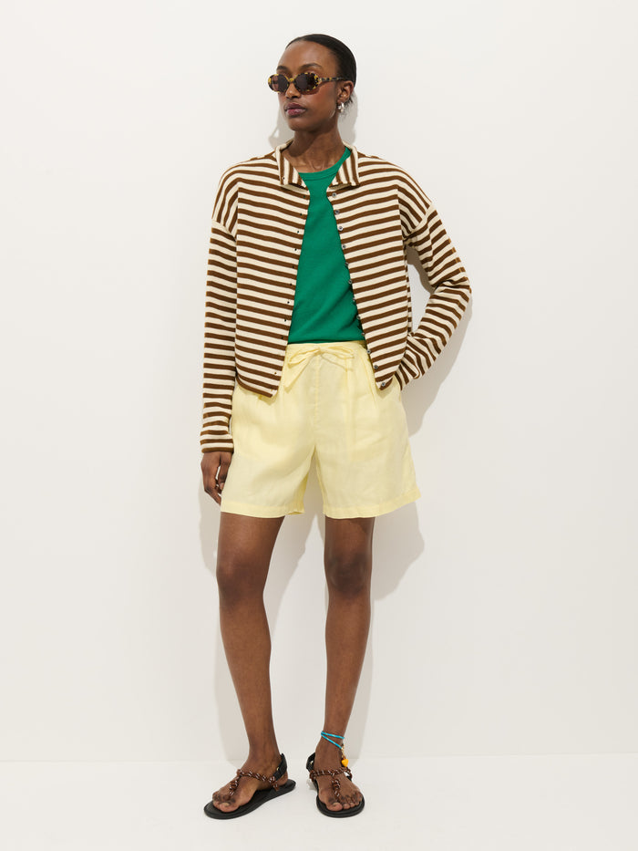 A person stands against a white background wearing sunglasses, the Taylor Striped Cardigan in Cotton Cashmere over a green shirt, yellow shorts, and sandals with an anklet. One hand is in their pocket.