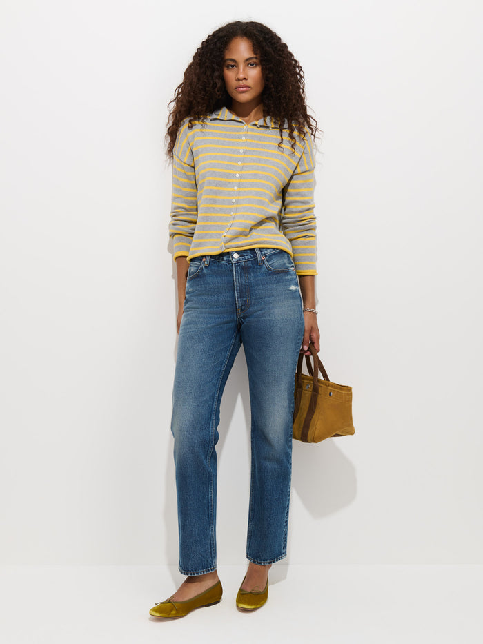 A woman with long curly hair stands against a white wall wearing the Taylor Striped Cardigan in Cotton Cashmere, paired with blue jeans, yellow flats, and holding a brown handbag.