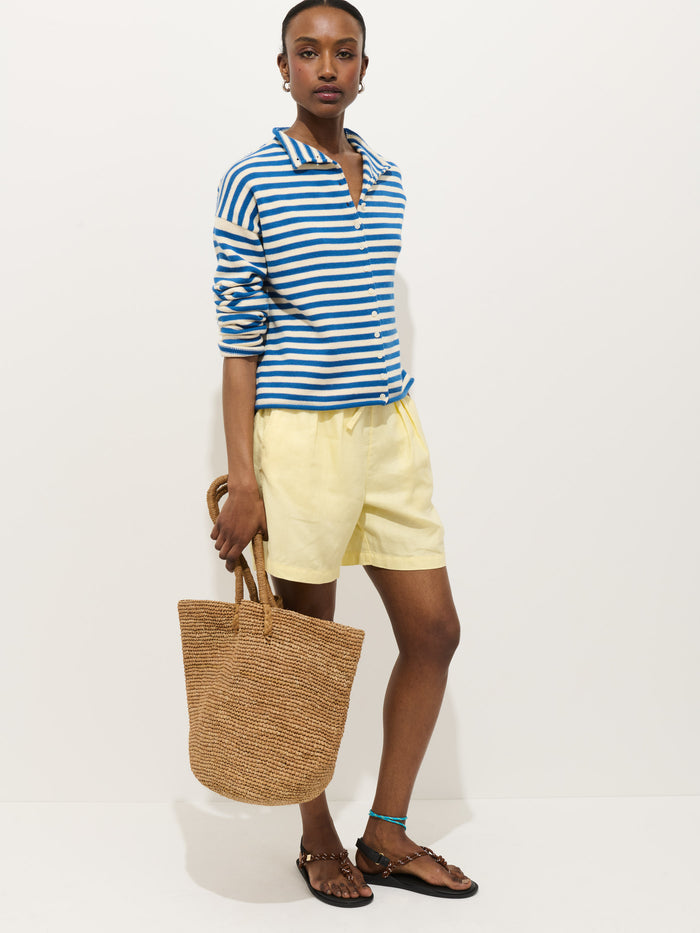 A person stands against a white background, wearing the Taylor Striped Cardigan in Cotton Cashmere—an oversized boxy blue and white striped top—paired with yellow shorts, black sandals, a woven straw tote bag, hoop earrings, and an anklet.