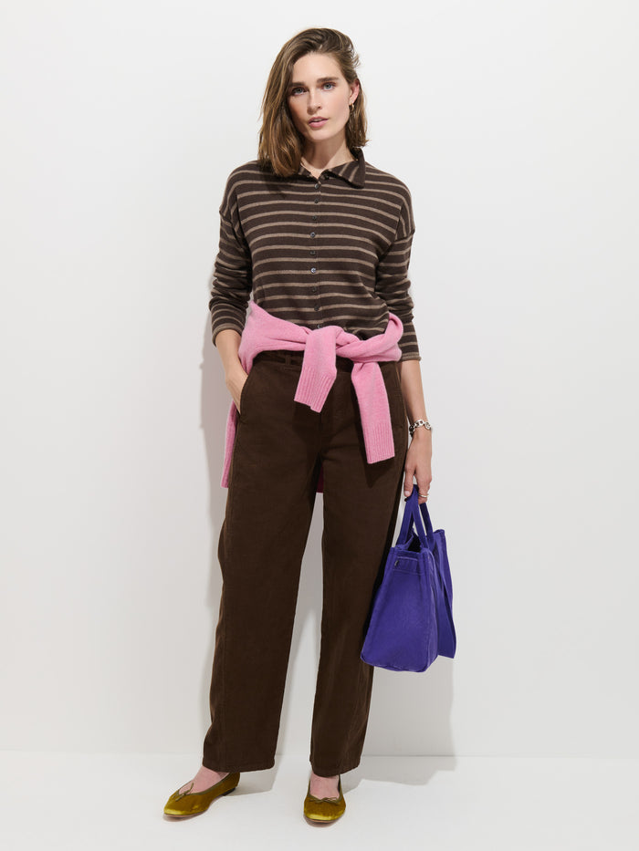 A woman stands against a white wall wearing the Taylor Striped Cardigan in Cotton Cashmere with troca shell buttons, paired with brown pants, yellow flats, a pink cotton cashmere sweater tied at her waist, and holding a blue handbag.