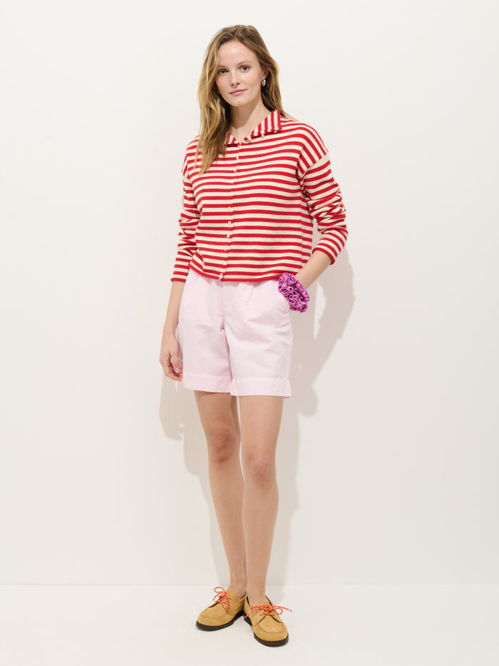 A woman stands against a white background wearing the Taylor Striped Cardigan in Cotton Cashmere, featuring an oversized boxy fit and red and white stripes, paired with light pink shorts, tan shoes with orange laces, and a purple scrunchie.