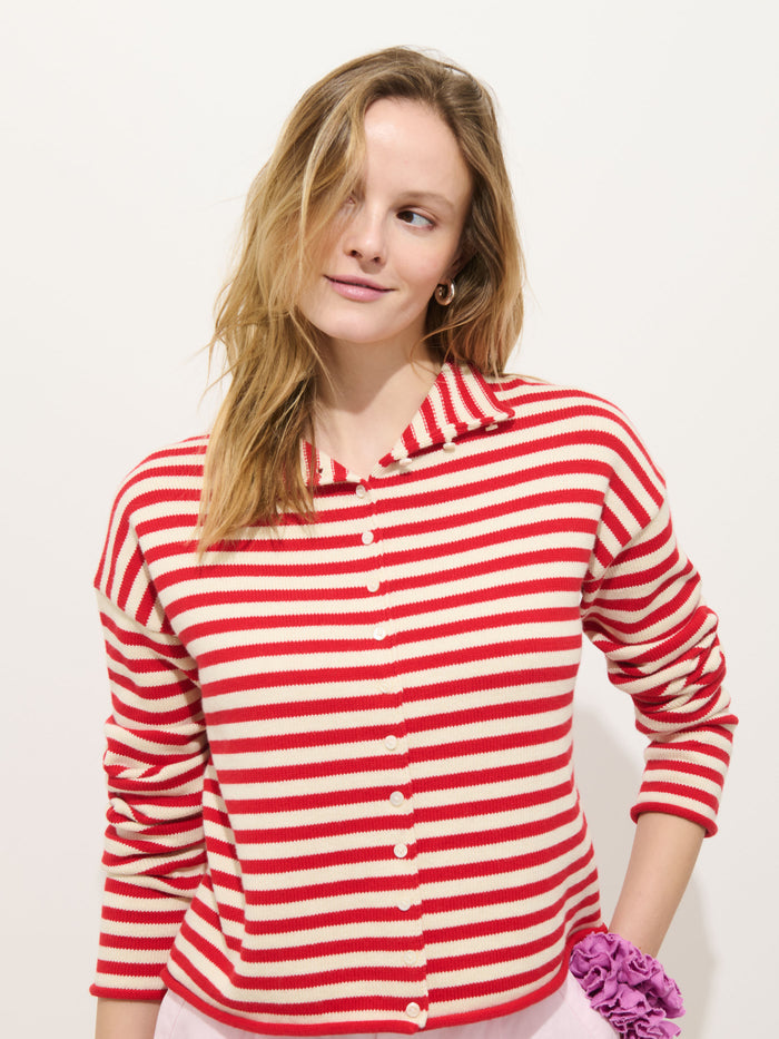 A woman with long blonde hair wears the Taylor Striped Cardigan in Cotton Cashmere with Troca shell buttons, paired with pink pants and a purple scrunchie on her wrist, standing against a plain white background and smiling slightly.