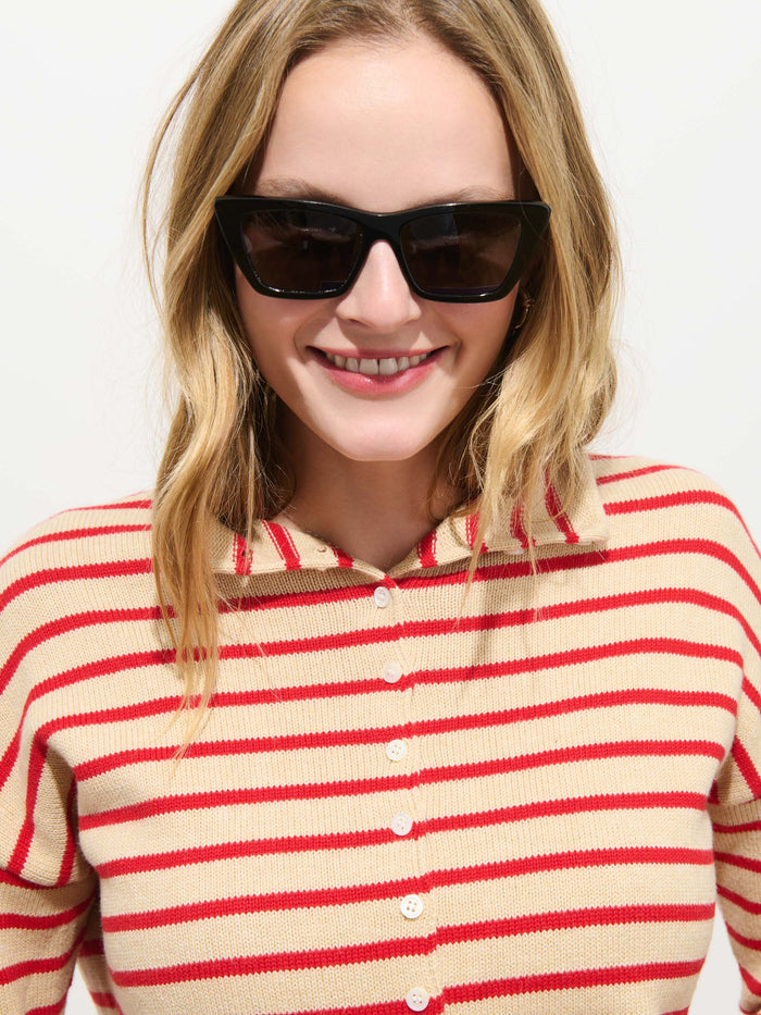A smiling person with wavy blonde hair wears black sunglasses and the Taylor Striped Cardigan in Cotton Cashmere, layered over a beige and red striped button-up shirt, standing in front of a plain white background.