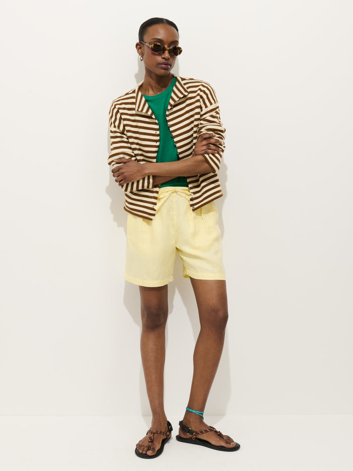 Person posing against a white background, wearing the Taylor Striped Cardigan In Cotton Cashmere—an oversized boxy fit brown and white striped jacket—over a green shirt, yellow shorts, brown sandals, sunglasses, and a blue anklet.