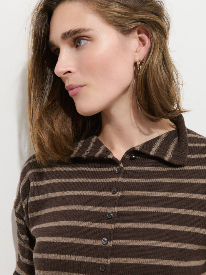 A person with light skin and shoulder-length brown hair wears the Taylor Striped Cardigan in Cotton Cashmere, featuring brown and beige stripes and Troca shell buttons, along with a small gold hoop earring, looking to the side against a plain white background.