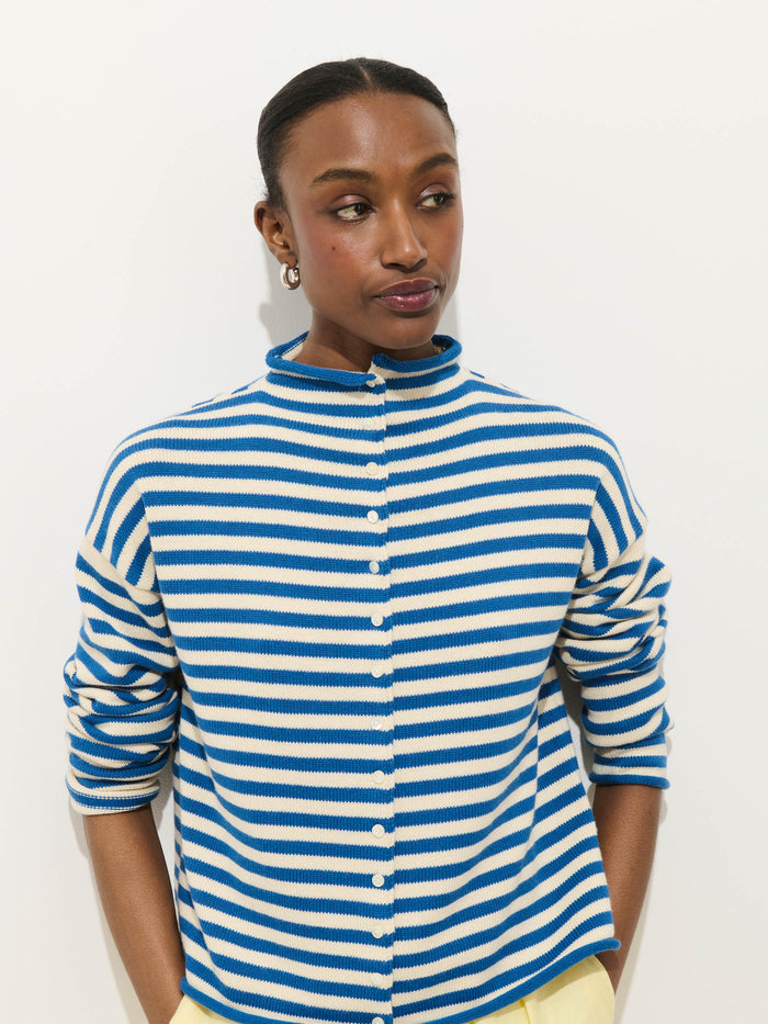 A woman with dark hair pulled back wears pearl earrings and the Taylor Striped Cardigan in Cotton Cashmere, featuring blue and white stripes. She stands against a plain white background, looking off to the side with a relaxed expression.