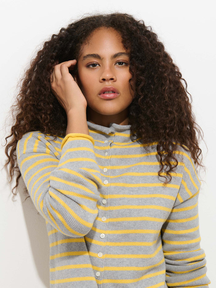 A person with long, curly brown hair wears the Taylor Striped Cardigan in Cotton Cashmere, featuring oversized gray and yellow stripes, standing against a white background and touching their hair with one hand.