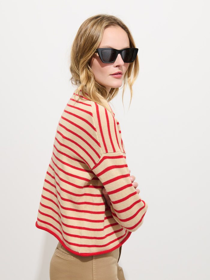 A woman in sunglasses wears the Taylor Striped Cardigan in Cotton Cashmere, featuring beige and red stripes, paired with tan pants as she stands against a plain white background, looking over her shoulder.