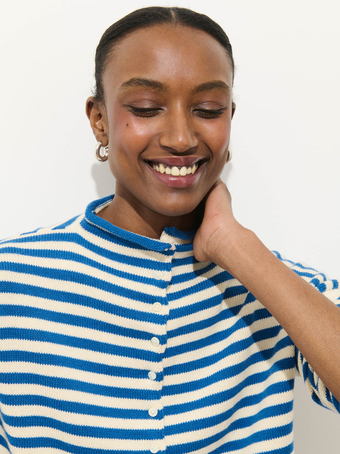 A person with dark skin and gold hoop earrings smiles with eyes closed, touching their neck. They wear the Taylor Striped Cardigan In Cotton Cashmere against a plain white background.