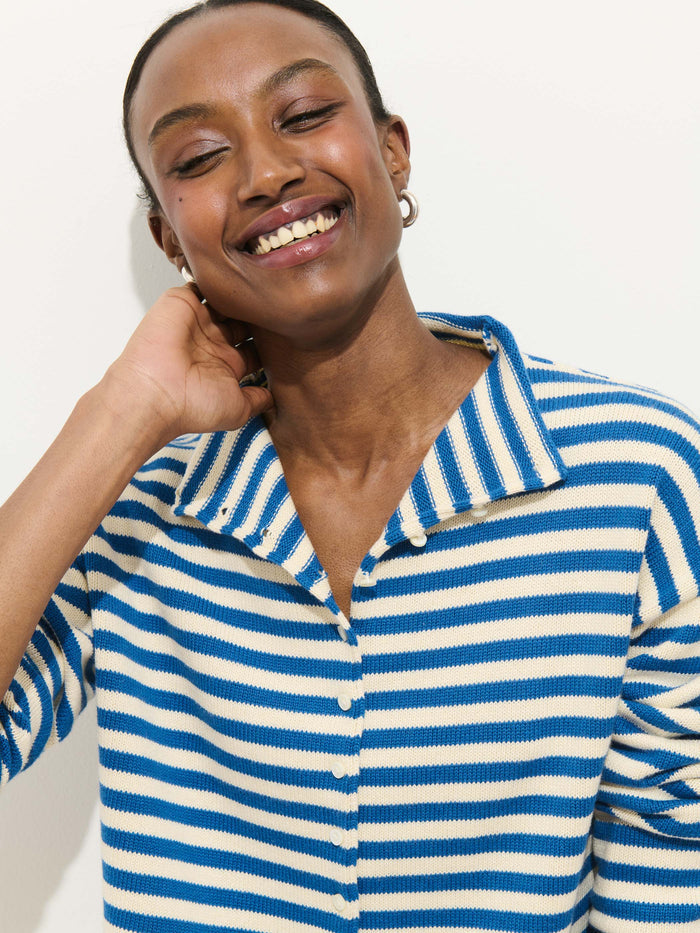 A person smiles with eyes closed, wearing the Taylor Striped Cardigan in cotton cashmere over a blue and white collared shirt, gold hoop earrings, and standing against a plain light background.