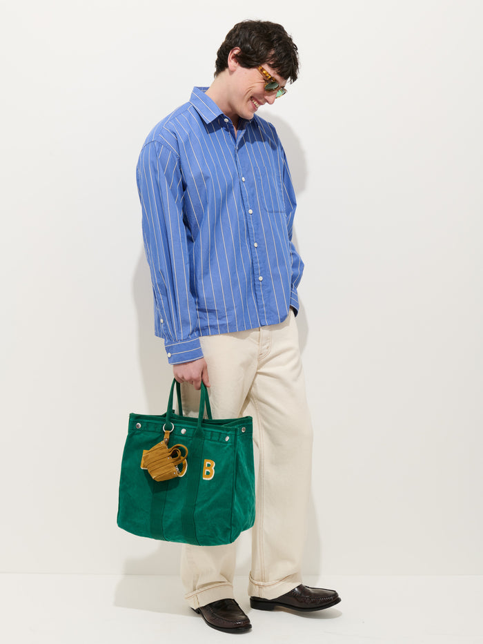 A person in a blue striped shirt, cream pants, and brown shoes stands against a white background, holding a large green tote bag with a yellow letter B and the Teeny Tiny Tote Keychain attached.