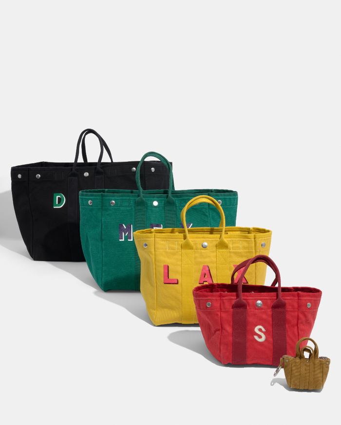 Five tote bags—black, green, yellow, red, and the Teeny Tiny Tote Keychain in brown—are lined up diagonally. Each has handles and side lettering; the smallest brown bag also functions as a keychain pouch with a handy clip.