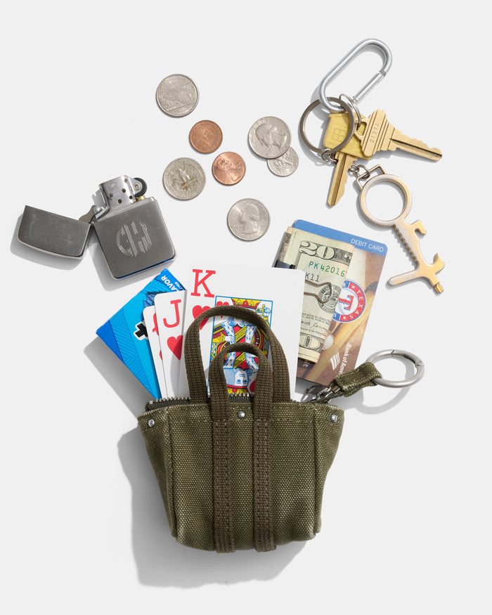 A small green Teeny Tiny Tote Keychain with playing cards, a carabiner, cash, a lighter, coins, keys, and multitools scattered around it—each casting shadows on a white background.