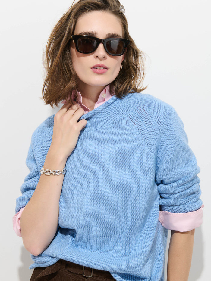 A woman with shoulder-length brown hair and dark sunglasses stands against a plain white background, wearing The Eastport Rollneck In Cotton—a light blue 100% cotton sweater—layered over a pink collared shirt and accessorized with a silver bracelet.