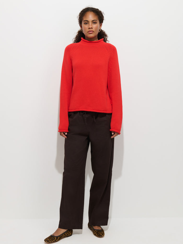 A person stands against a white background wearing The Eastport Rollneck in Cotton in bright red, loose dark brown pants, and leopard print flats.