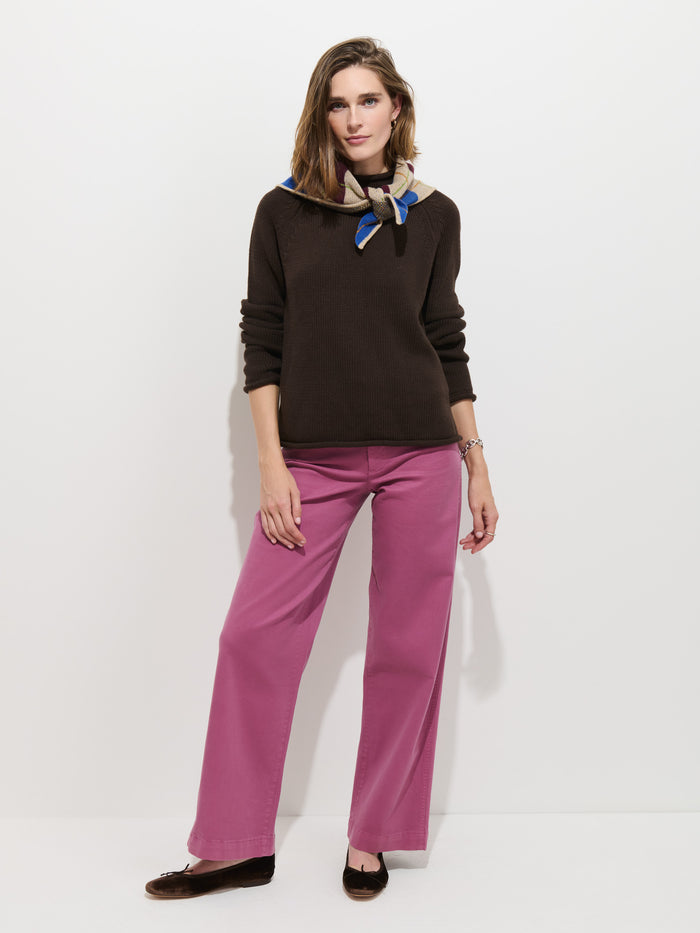 A woman stands against a white wall, wearing The Eastport Rollneck In Cotton—a dark roll-neck sweater—paired with a blue patterned scarf, wide-leg pink pants, and black shoes. She has one hand relaxed by her side and the other in her pocket.