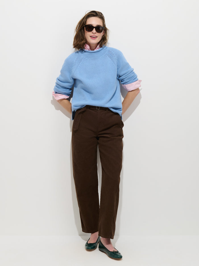 A person stands against a white wall wearing sunglasses, The Eastport Rollneck In Cotton in light blue over a pink shirt, brown pants, and green flats, smiling with hands in their pockets.
