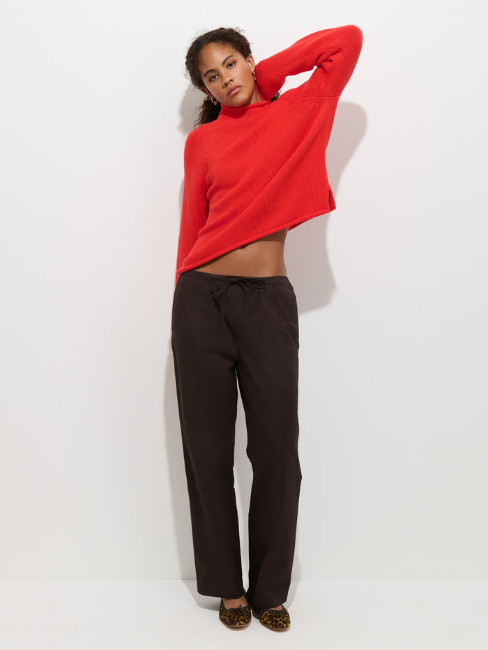 A person stands against a white wall, wearing The Eastport Rollneck In Cotton in bright red, paired with black pants and leopard print flats, with one arm raised behind their head and the other hanging by their side.