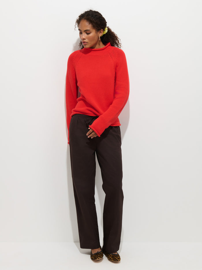 A woman stands against a white wall wearing The Eastport Rollneck In Cotton—a bright red sweater—paired with loose dark brown pants and leopard print shoes. She looks down and to the side, with a yellow flower tucked behind her ear.