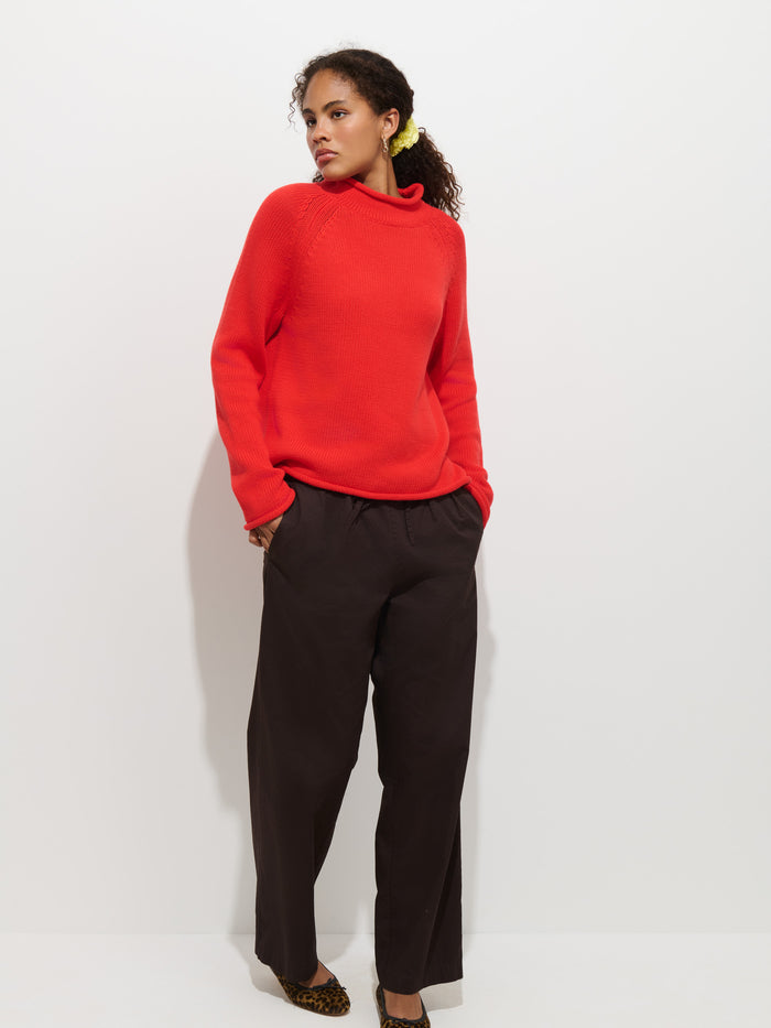 Wearing The Eastport Rollneck In Cotton in bright red, paired with loose dark brown pants and leopard print shoes, a person stands against a plain white background, gazing to the side with one hand in their pocket.