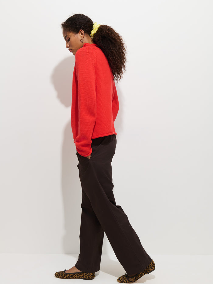 A person with curly hair in a yellow scrunchie wears The Eastport Rollneck In Cotton—a cozy, red 100% cotton sweater—paired with black pants and leopard print flats while walking sideways against a plain white background.