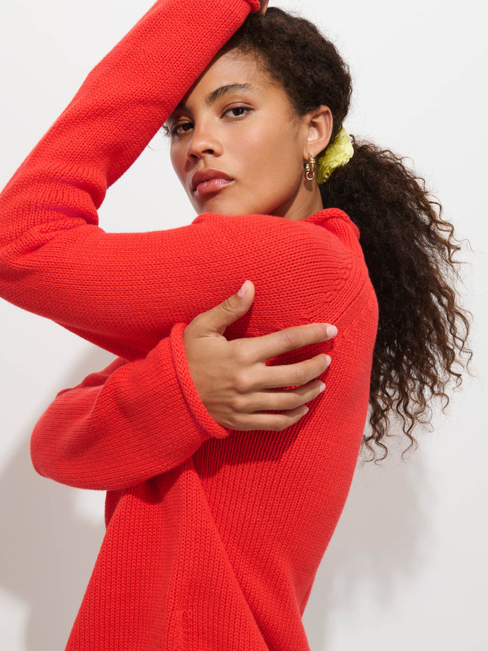 Wearing The Eastport Rollneck In Cotton, a woman with curly hair styled in a yellow scrunchie poses confidently against a plain white background—one arm raised, the other wrapped across her chest—showcasing the bright red, 100% cotton sweater.