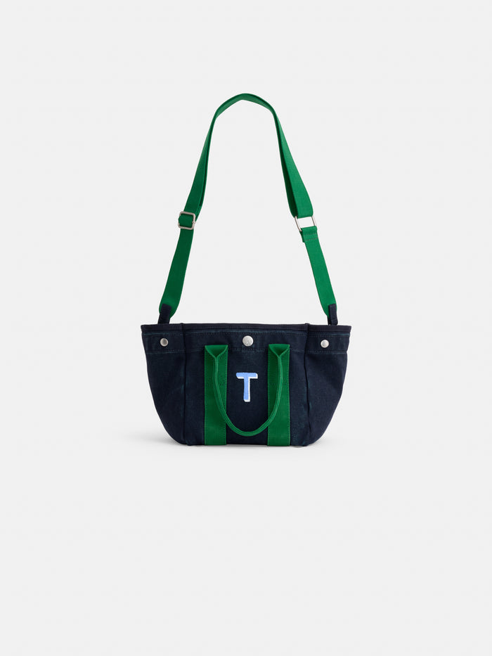 The Mini is a black cotton canvas mini tote with green straps and a wide, adjustable shoulder strap. The front has a blue patch displaying a white letter T for a stylish personalized touch. Photographed on a plain white background.