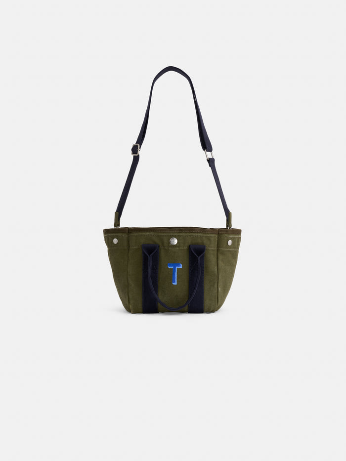 The Mini: An olive green cotton canvas mini tote with dark blue handles, a blue 'T' on the front, shoulder strap, and silver hardware, shown against a plain white background.