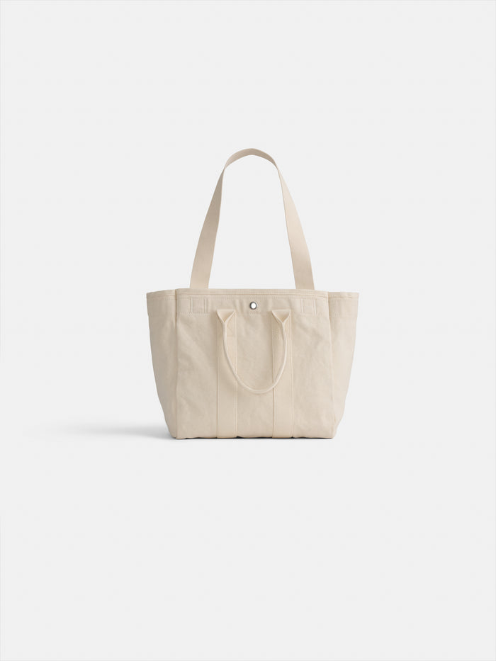 The Perfect Daily Tote, made of beige cotton, features two short handles, two long shoulder straps, and a simple snap closure at the top center. Displayed against a plain white background.
