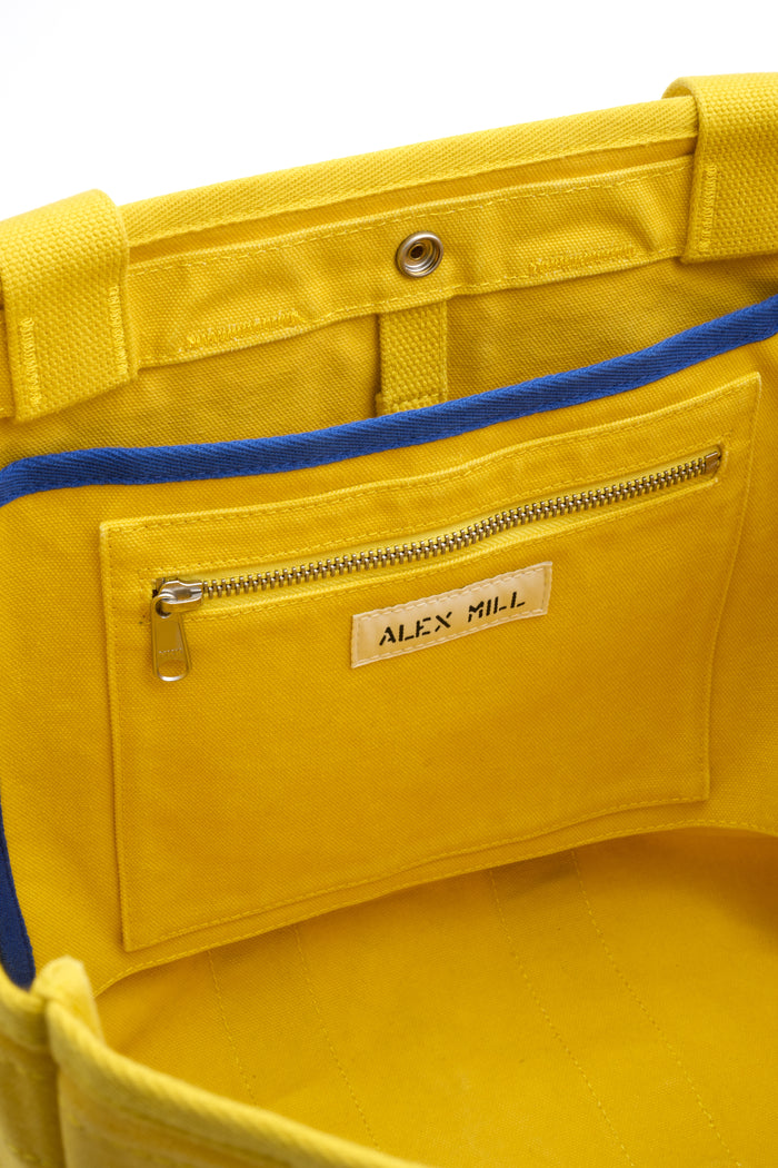 Close-up of the inside of The Perfect Weekday Tote, a yellow cotton bag with blue trim, showing a zippered pocket and a label reading ALEX MILL.