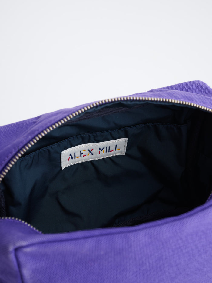 A close-up of an open purple The Perfect Dopp Kit reveals a dark-lined interior and an interior label that reads ALEX MILL in colorful letters.