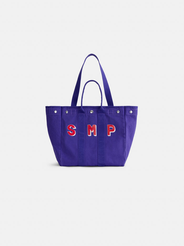 Introducing the Painted Perfect Weekday Tote—a purple bag with two handles and silver rivets, adorned with bold red and white “SMP” lettering on the front. This stylish accessory is perfect for anyone who loves personalized totes and monogrammable bags.