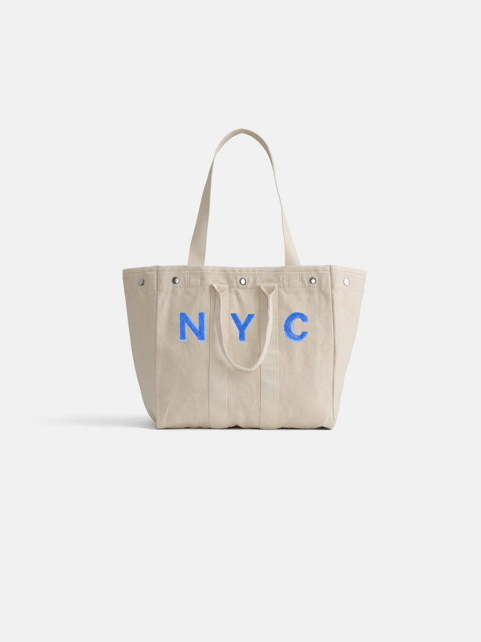 Introducing The Perfect Weekday Tote: crafted from beige cotton with sturdy double handles, this chic accessory showcases bold NYC lettering in blue across the front and is finished with silver rivets at the top corners. Ideal for daily use, it’s also part of our monogrammable tote bag collection—so you can personalize it to make it truly one-of-a-kind.