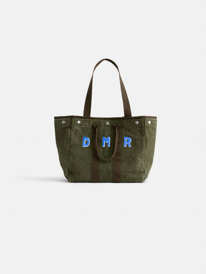 The Perfect Weekday Tote is a green suede bag with brown handles, featuring large blue and white "DMR" letters monogrammed on the front, displayed against a plain white background.