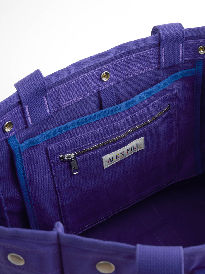 Close-up of the interior of The Perfect Weekday Tote in purple fabric, showcasing a zippered pocket with blue trim and a label marked “ALEX MILL.” This tote features snap closures, sturdy handles, and an elegant design—ideal for those looking for monogrammable bags or a personalized tote bag option.