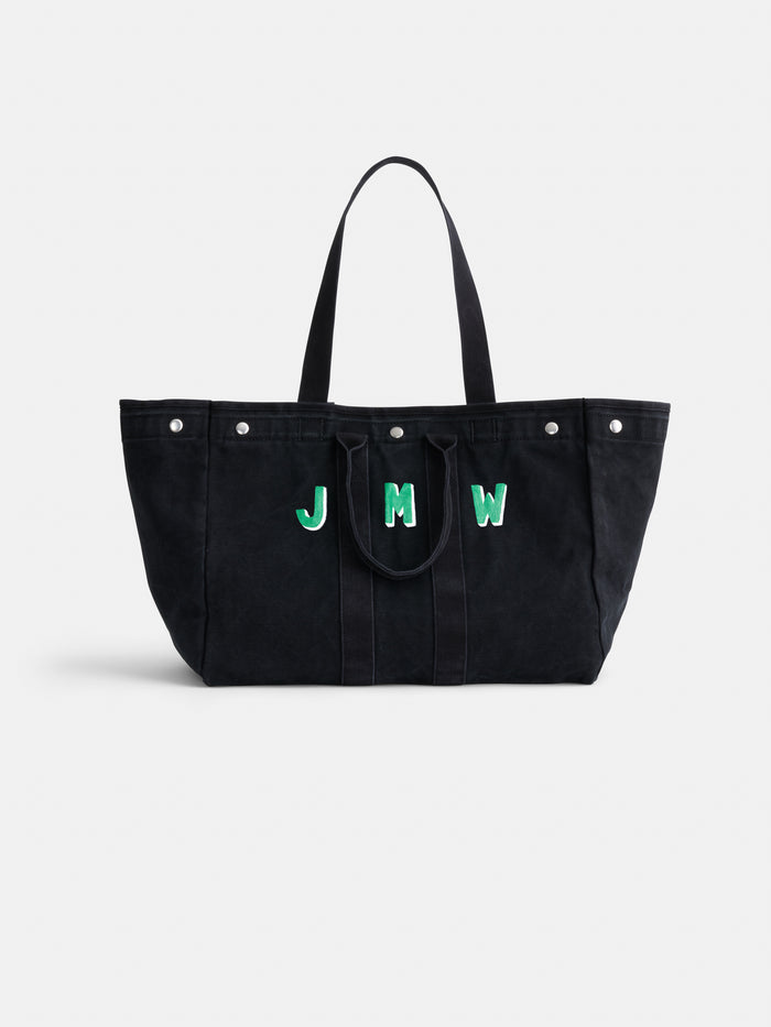 The Perfect Weekend Tote is a large black cotton bag with two handles, silver studs, and a monogram of the letters JMW in green-bordered white font on the front. This personalized tote is displayed against a plain white background.