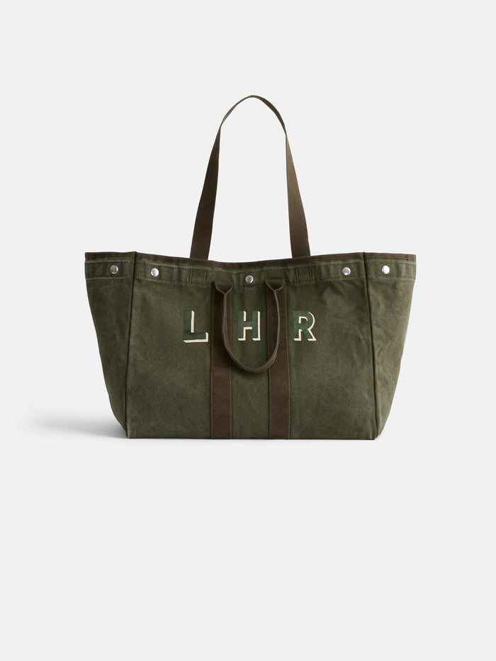 The Perfect Weekend Tote is a large olive green cotton bag with brown handles and trim, featuring "LHR" in white and green on the front. It includes personalized details and metal snaps near the top edges.