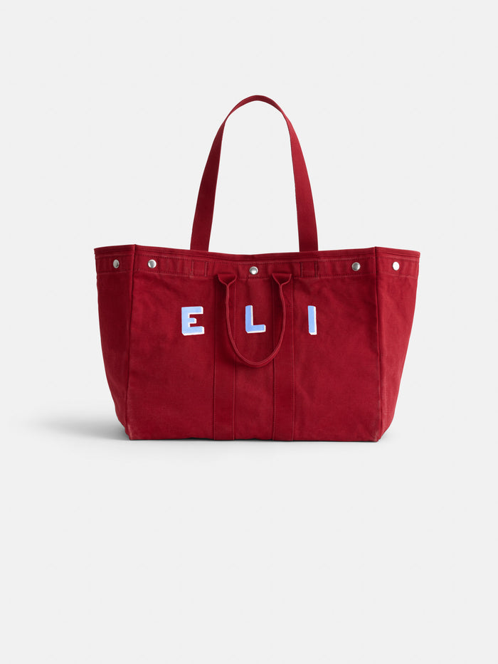 The Perfect Weekend Tote is a large red cotton bag with two handles and ELI in bold white and blue on the front. It features metal rivet accents, monogram options, and a simple, structured design for everyday style.