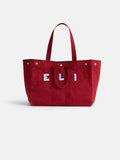 The Perfect Weekend Tote is a large red cotton bag with two handles and ELI in bold white and blue on the front. It features metal rivet accents, monogram options, and a simple, structured design for everyday style.