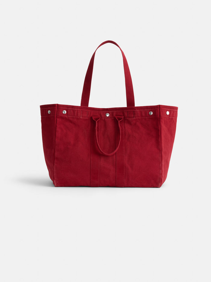 The Perfect Weekend Tote is a large, deep red cotton bag with two short handles, silver rivets, and a plain design, ideal for classic monogramming lovers. Shown against a white background.
